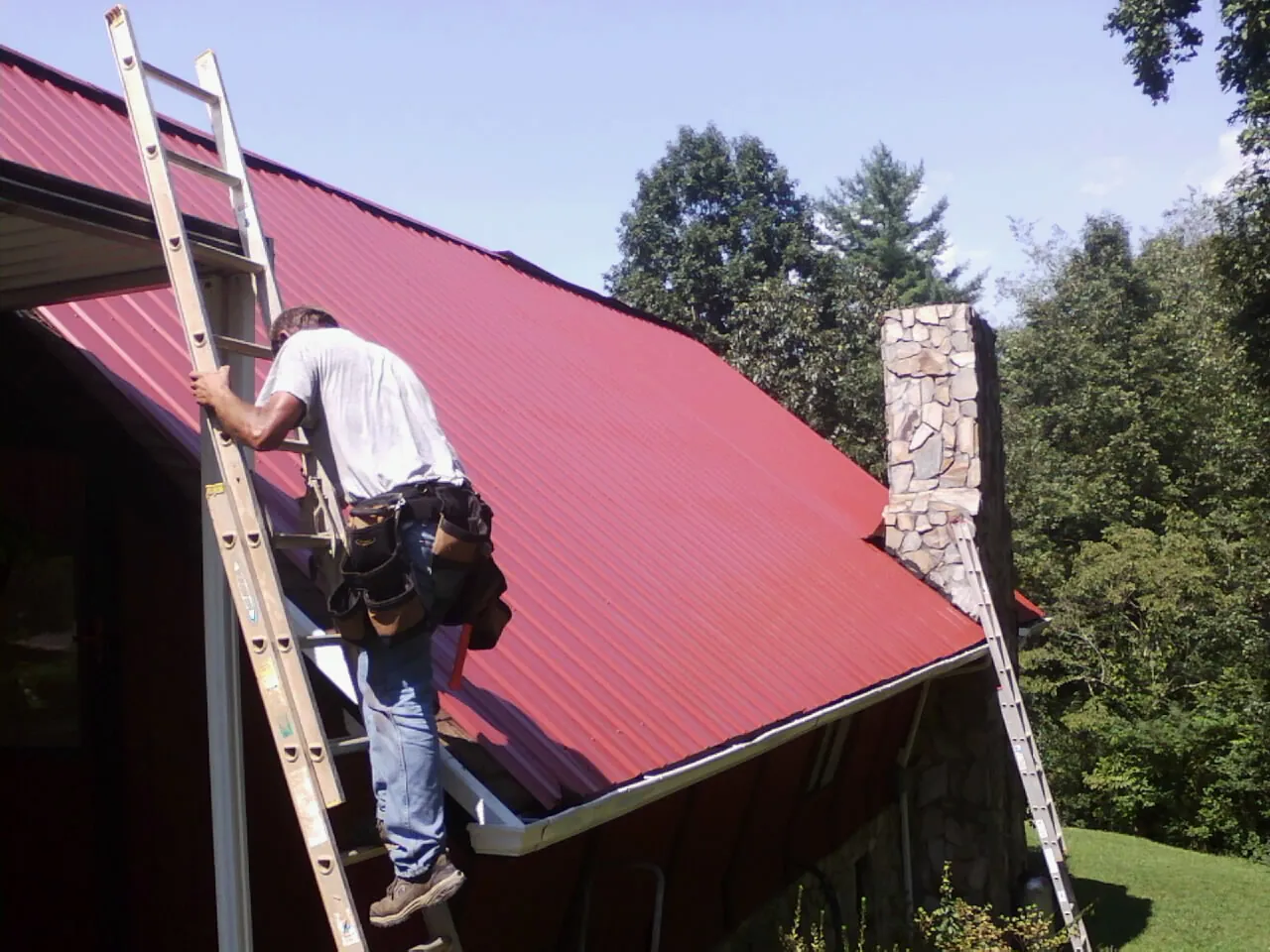 Roofer on a ladder beside a red metal roof and stone chimney — Soffit & Fascia Repair by Roofing Brothers in Anvik